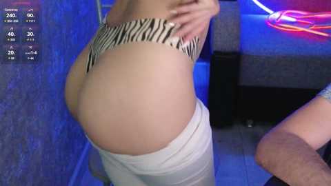 A video shows a woman's lower body in a tight, zebra-print thong, highlighting her round buttocks. She wears white pants and a purple top. Background features blue and pink neon lights.