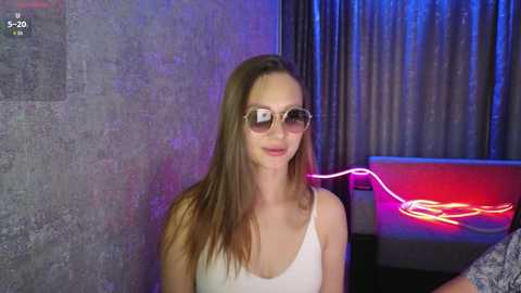 Video of a young woman with long brown hair, wearing sunglasses and a white tank top, standing against a textured gray wall with a red neon sign and dark curtains in the background.
