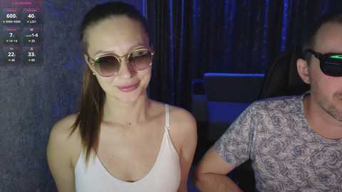 Video of a fair-skinned woman with brown hair, wearing sunglasses, a white tank top, and a subtle smile, standing next to a man in a patterned shirt, in a dimly lit room.