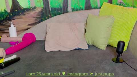 Media: Video of a colorful, themed bedroom with a green forest mural, featuring a beige pillow, a neon yellow pillow, a pink vibrator, and a black microphone on a gray carpet.