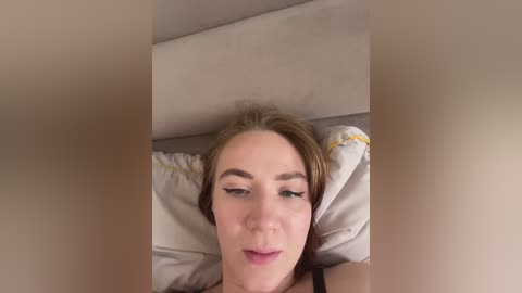 A video of a light-skinned woman with blonde hair, lying on a bed with beige pillows, wearing a black top, smiling subtly. The image has a soft focus and is taken from above.