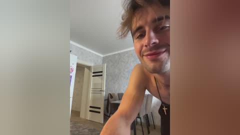 A video of a shirtless young man with messy hair and a friendly grin, captured from a low angle in a dimly lit, cluttered room with beige walls and a fridge in the background.