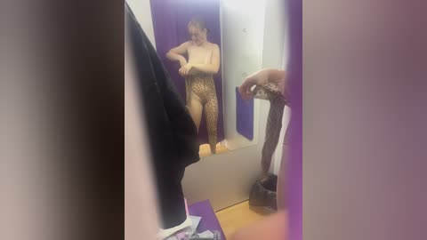 A blurry, dimly-lit video of a woman in a leopard print bodysuit, standing in a dressing room, with a mirror reflecting her partially. The background shows purple and white walls, a black coat, and scattered clothes on the floor.