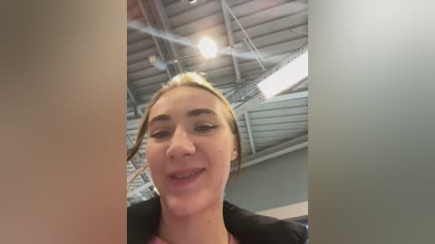 Video of a young woman with fair skin, light brown hair in a bun, and black clothing, smiling. Background features a ceiling with exposed beams and fluorescent lights, giving a modern, industrial vibe.