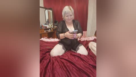 A middle-aged woman with short, platinum blonde hair, wearing a black lace robe and white stockings, sits on a bed with red velvet sheets, absorbed in her smartphone. The background shows a cluttered dresser with personal items.