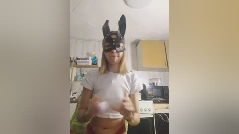 Media: A video of a woman with light skin, wearing a black bunny mask, a white crop top, and red shorts, standing in a kitchen with white cabinets, a stove, and a microwave.