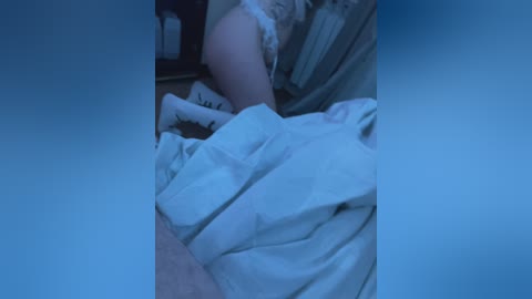 A dimly lit, blurry video shows a person lying in bed, partially covered by white sheets. The background includes a dark wall and indistinct objects. The scene has a blue tint, suggesting a nighttime setting.