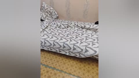 Video of a bed with a white comforter featuring a repeating grey heart pattern, against a beige wall and a yellow patterned carpet. The image is slightly blurry.