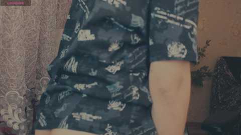 Video of a person wearing a dark blue, short-sleeved shirt with white space-themed designs, partially visible from the waist up, in a room with a lace curtain and a partially visible plant.