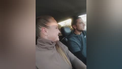 Video of a woman with blonde hair tied back, wearing a light brown turtleneck, and a man in a black jacket sitting in a car.