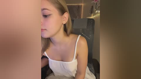 Media: Video of a young, fair-skinned woman with straight, light brown hair, wearing a white spaghetti-strap top, looking contemplative in a dimly lit room with fairy lights and a grey chair.