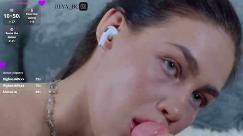 Video of a young woman with fair skin, brown hair tied back, wearing an earbud and a silver necklace, performing oral sex on a man, in a dimly lit room.