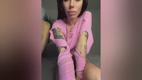 Media: Video of a fair-skinned woman with long brown hair, wearing a pink, ribbed sweater with lace-up front, revealing tattoos on her arms, sitting on a gray couch in a dimly lit room.