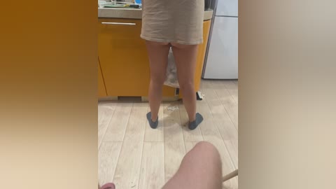 A video of a woman in a beige dress and blue socks standing in a kitchen with light wooden floors and orange cabinets.
