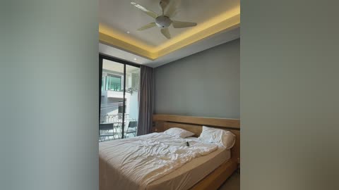 Media: Video of a modern bedroom with a wooden bed, white linens, a ceiling fan, and large glass sliding doors leading to a balcony. Gray walls and soft lighting create a serene ambiance.