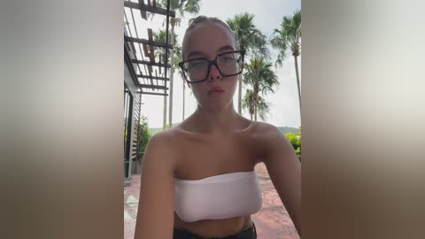 Media: A video of a young woman with light skin and brown hair tied back, wearing black-framed glasses and a white strapless bandeau top. She stands in a tropical outdoor setting with palm trees and a metal structure in the background.