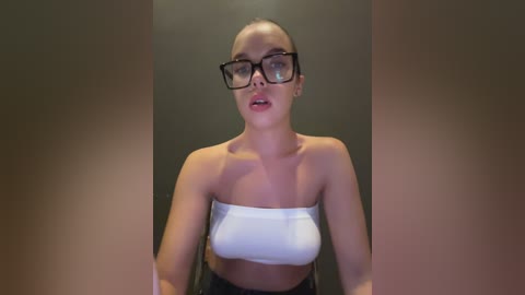 Media: Video of a fair-skinned woman with short hair, wearing black-rimmed glasses, a white bandeau top, and black pants. She stands against a plain, dark background.