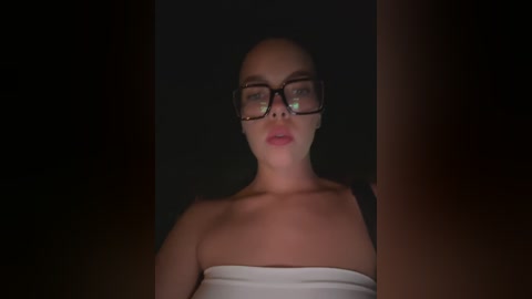 Media: Video of a bald woman with glasses and a white strapless top, standing in a dimly lit room. Her expression is neutral, and the background is dark and indistinct.