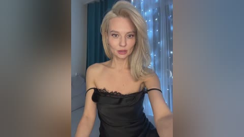 Video of a fair-skinned, blonde woman with straight hair, wearing a black, off-shoulder satin dress, standing indoors against a backdrop of blue curtains and fairy lights.