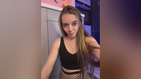 Media: Video of a young Caucasian woman with long, straight blonde hair, wearing a black crop top and fishnet stockings, standing in a dimly lit, modern kitchen with pink and purple accents.