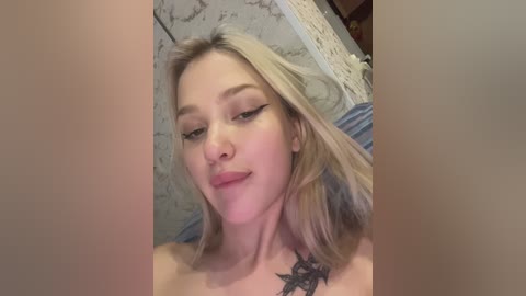 Media: A video of a fair-skinned, blonde woman with shoulder-length hair and a black tattoo on her chest, wearing eyeliner. She has a confident expression and is topless. The background shows a marble wall and a partially visible bed.