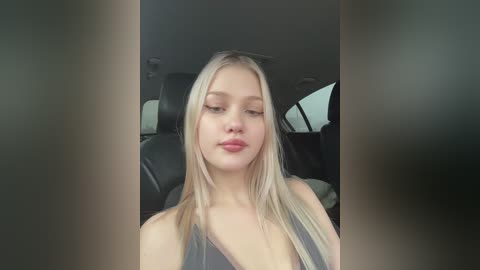 Media: Video of a young, fair-skinned woman with long, straight blonde hair, wearing a black sleeveless top, sitting in a car with dark interior. She has a neutral expression.