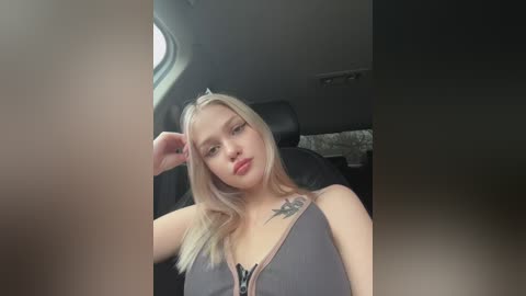 Media: Video of a blonde woman with fair skin, wearing a gray tank top, sitting in a car. She has a tattoo on her left shoulder. The background shows the car's interior.