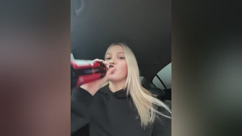 Media: Video of a blonde woman in a black coat, drinking from a red can inside a car. The background is blurred, with a dark, possibly grey, interior.