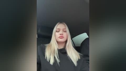 Media: Video of a young blonde woman with long hair, wearing black, sitting in a car with closed eyes and a serene expression.