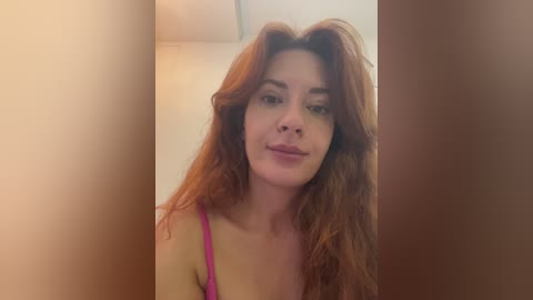 Media: Video of a young woman with long, wavy red hair and fair skin, wearing a pink tank top, standing in a dimly lit room with beige walls.