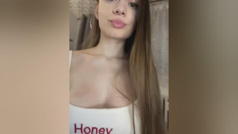 A video of a young woman with light skin and long brown hair, puckering her lips. She's wearing a white, low-cut top with \"Honey\" written in red. The background shows beige walls and a window with blinds.