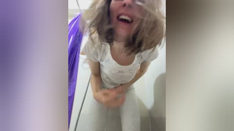 A video shows a woman with shoulder-length blonde hair, wearing a white t-shirt and light pants, laughing while sitting on a toilet, reflected in a glass door.