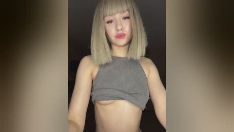 Media: Video of a young woman with pale skin, straight platinum blonde bob haircut, wearing a cropped gray top, exposing her small breasts. Background is blurred, featuring neutral tones.