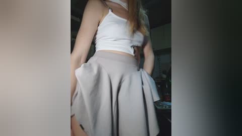 A video of a slender woman in a white crop top and high-waisted, light gray pleated skirt, standing indoors with dim lighting.