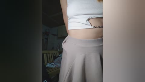 Video of a young woman's midsection, showing a white crop top and light beige high-waisted skirt, in a dimly lit, cluttered room with a wooden chair and scattered clothes.