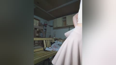 Video of a cluttered, dimly lit workshop with a person wearing a white dress and holding a wooden plank, amidst various tools and wooden boards.