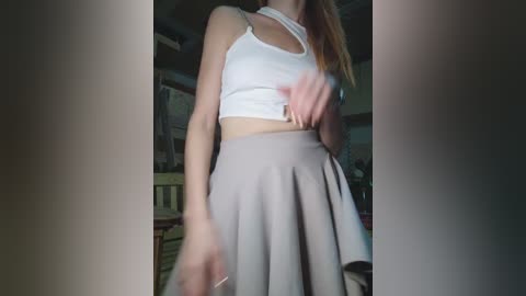 Video of a woman with light skin and straight, long blonde hair, wearing a white crop top and a grey, pleated skirt, standing indoors in a dimly lit room.