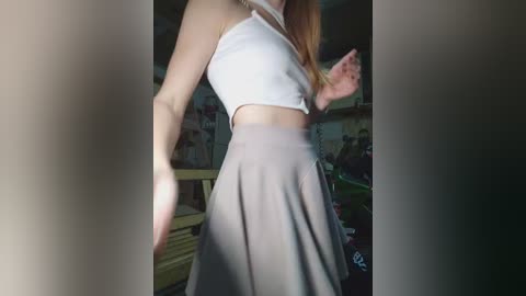 Video of a slim, light-skinned woman with long, straight, light brown hair, wearing a white crop top and high-waisted, light gray skirt, captured in a dimly lit room with cluttered shelves and furniture.