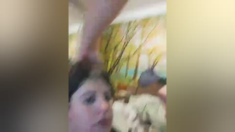 A blurry video shows a young woman with fair skin and dark hair, partially obscured by a hand. The background features a colorful, abstract painting with green, yellow, and orange hues. The image is grainy and out of focus.