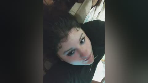 A close-up video of a young woman with curly dark hair, wearing a black shirt, lying on a bed with beige sheets and pillows. The image is dimly lit, giving a soft, intimate atmosphere.