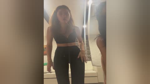Video of a slender, light-skinned woman with long brown hair in a dark bra and high-waisted pants, standing in a dimly lit bathroom.