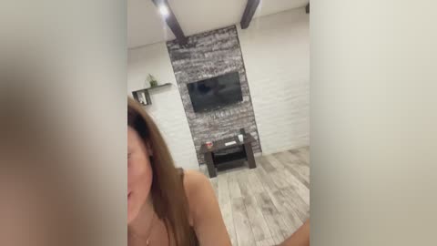 Video of a modern living room with a young woman with long brown hair, wearing a top, standing in front of a black-and-white tiled fireplace.