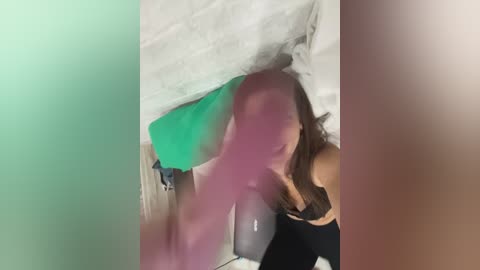 Video of a woman with light skin, long brown hair, wearing a black bra, partially obscured by a green and white blanket. Background shows a white wall with a towel hanging.