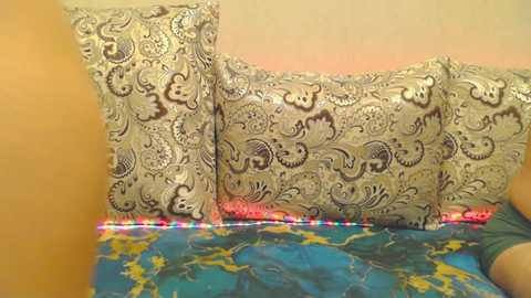 Media: A video of a bed with a turquoise and yellow map-patterned duvet. The background features a beige wall. The bed is adorned with three large, ornate silver pillows with intricate, floral patterns.