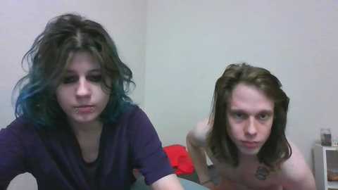 Media: A video of two young women with messy hair, one with blue-streaked brown hair and the other with long brown hair. They are topless, wearing only black shirts, in a dimly lit room with a red blanket and a white shelf in the background.