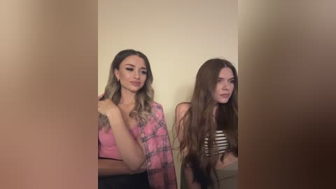 Media: Video of two young women with long hair, one in a pink plaid shirt and the other in a striped top, standing against a beige wall.