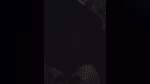 A dimly lit video of a person with dark hair, wearing a mesh mask, looking upwards, taken from a low angle. The background is mostly black, emphasizing the shadowy figure.