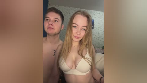 Video of a young, fair-skinned couple, both shirtless. The man has short brown hair and a small tattoo, while the woman, with long blonde hair, wears a white bra. Background shows a blue wall and floral-patterned wallpaper.