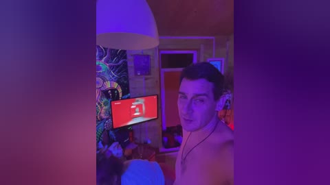 Video of a man with short dark hair, wearing headphones, standing in a dimly lit room with purple lighting, holding a phone showing a red screen. The background features colorful tapestries and a hanging lamp.