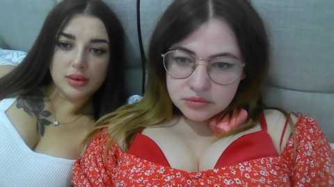 Video of two young women with fair skin, one with long dark hair and glasses, the other with shoulder-length hair, both wearing red tops, sitting on a gray couch.
